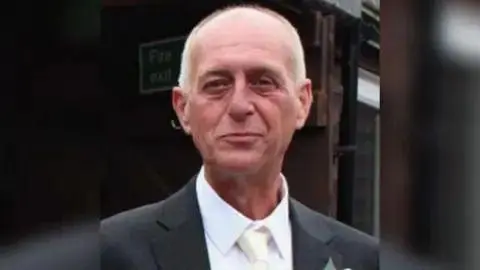 Kent Police Alan Tomkins looks at the camera and is smiling. He is wearing a dark suit with a white tie and white shirt.