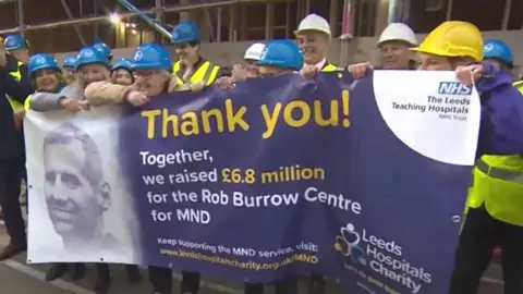 Smiling people in hard hats unfurl a banner which reads: "Thank you! Together we raised £6.8million for the Rob Burrow Centre for MND." Rob Burrow's face is pictured in black and white on the left-hand side of the photo. 