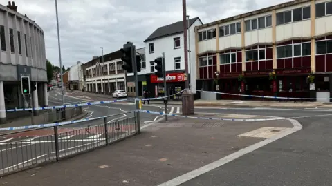 A police cordon in place on a high street in a city centre. The cordon is across a set of traffic lights and blocks multiple roads.