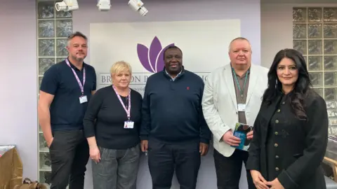 Shariqua Ahmed/BBC Staff from the Bretton practice - Jason wearing a blue T-shirt, Jackie wearing a black jumper and a purple lanyard, practice partner Dr Mark in a navy blue zipped jumper, councillor Richard in a white blazer and Dr Shabina in a black coat with gold buttons.