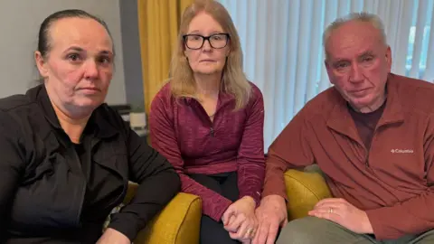 A woman in a black top on the left sitting on a mustard coloured sofa, next to a woman with blonde hair and a pink long-sleeved running top on, who is next to a man with a red long-sleeved jumper on. All look at the camera with a sad and concerned look on their faces.