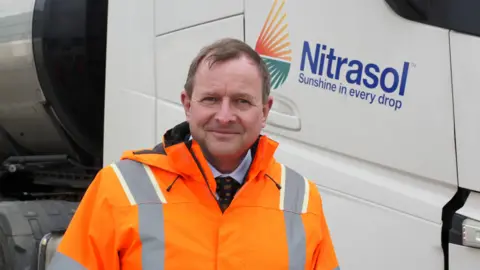 John Fairhall/BBC A man in a hi-vis jacket standing in front of a white lorry cab with "Nitrasol" branding on it.