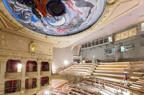 Anneleen Lindsay A huge blue mural on the roof of the theatre which features a moon and clouds underneath rows of half constructed seats can be seen as scaffolding towers in front of the stalls on the left 