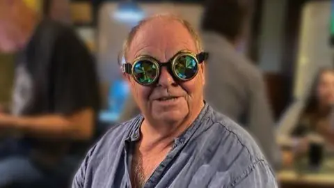 Contributed Lawrence Dixon is wearing a blue shirt and is wearing goggles that cover both eyes and has a kaleidoscope like pattern on them. There are several people sitting behind him. 