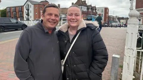 Lili Sheppard/BBC A man in a grey jumper and a woman in a black puffer jacket. They are standing at a High Street.