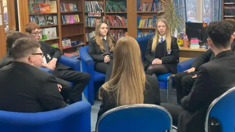 A group of children sit in the library at their school in a discussion group 