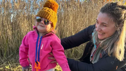 Handout Effie (left), a young girl, and her mum Rachel standing by a bush of rushes. Effie is wearing an orange winter bobble hat and a pink zip up jacket with sunglasses. Rachel is wearing a dark outdoor jacket and has long blonde hair.