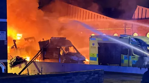 PA Media Large plumes of smoke rise from the wreckage of a burnt out ambulance. The thick smoke appears deep orange from the bright flames. The blackened hollow of one ambulance is being sprayed with water from hoses.