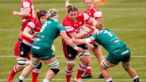 Getty Images Gloucester-Hartpury women's rugby players in red uniform playing against Trailfinders who are in green uniform.