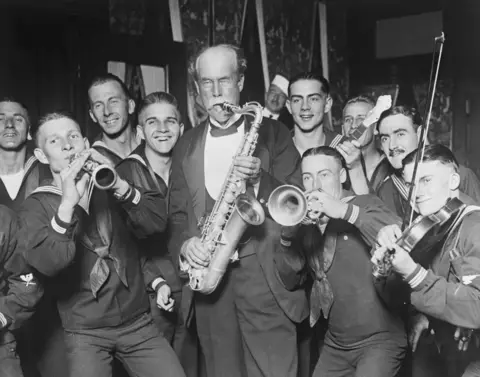 Bettmann/Getty A man with a moustache, wearing a dinner jacket and playing a saxophone, surrounded by musicians dressed as sailors