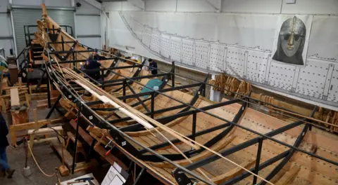 Jo Black/BBC A view of the longship in the process of being built inside a large warehouse. Planks of wood lie either side of the ship. There is an image of a silver helmet above the ship. 