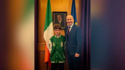 Boy, 13 with brown hair in a green jacket standing beside taller man in blue suit and red tie. They are standing in front of an Irish flag and a European flag.