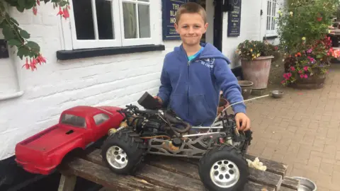 Hertfordshire Police A child stands outside a building, holding a large remote‑controlled off‑road toy vehicle placed on a wooden table.