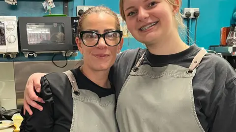 Jonny Humphries/BBC Natalie Attia, 42, who has dark brown hair tied back an is wearing large black framed glasses, a black top and a dark green apron, stands next to her colleague, who is dressed in similar clothing but has blonde hair and is a few inches taller, in the kitchen of a cafe. 
