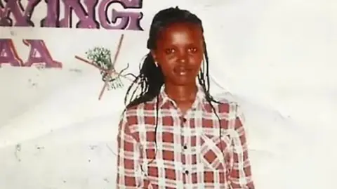 Wanjiru Family A full-body photo of Agnes Wanjiru wearing jeans and a chequered shirt