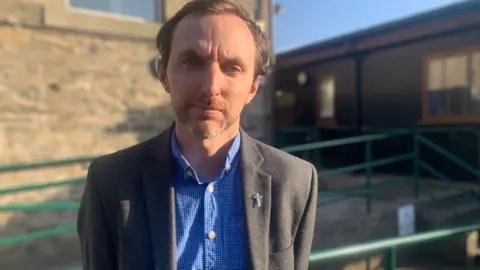 Nick Turley looking into the camera in front of the school. He has short, dark hair and is wearing a grey blazer with a blue checked shirt. 