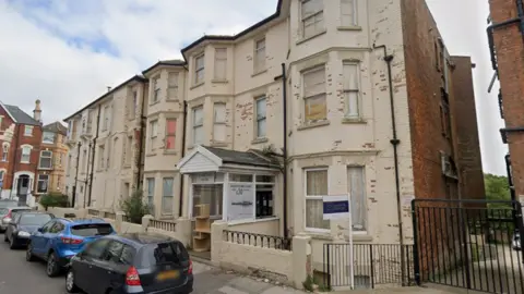 A three-storey white building with cars parked outside it. 