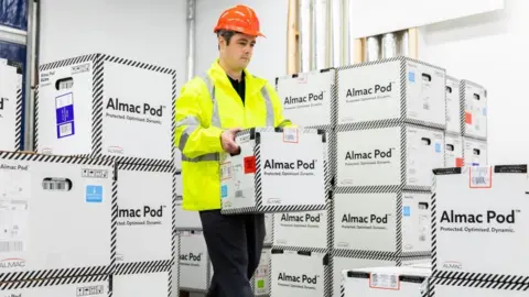 Almac An Almac worker wearing a fluorescent jacket and a a hard hat carries a box containing a product made by the company