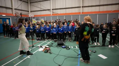 Mark Surridge A school gymnasium with green floors and white, red and blue sports markings. Ed Sheeran is standing in the foreground with his back to the camera. He has orange hair and is wearing a black top with multi-coloured palm trees on the back. Another adult is wearing a green vest with white trousers and dark-coloured boots. In the background is a choir of children. Most in the front row are wearing blue jumpers and dark trousers.