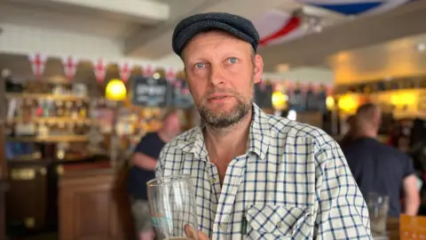 George Carden/BBC Graham Powell wearing a flat cap and chequered shirt and holding a pint of beer in the pub