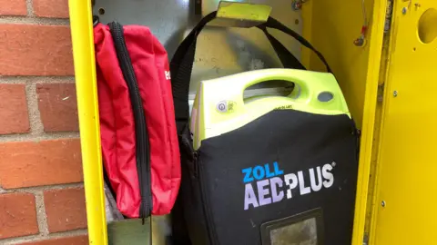 An open yellow defibrillator cabinet on a wall, showing a black defibrillator inside, with a red bleed kit next to it