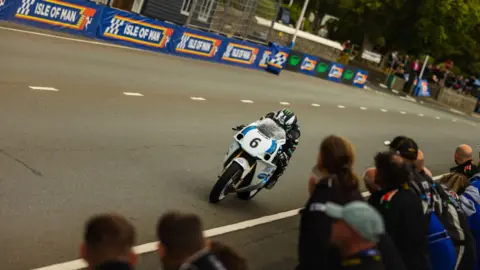 Classic TT Michael Dunlop, wearing mainly black leathers and a black-and-white helmet with a bright green logo, rides his motorbike on Glencrutchery Road with spectators looking on.