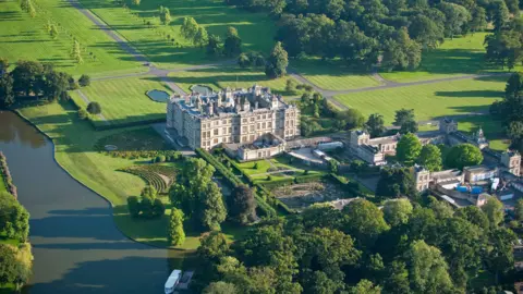 Longleat Drone shot of Longleat house and gardens with river running by on a sunny day. Lots of lush green lawns and trees.