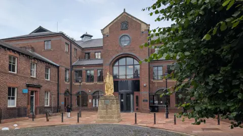 To the right in the foreground there is part of a green tree. In the centre there is a plinth with a yellowing statue. In the background there is a red brick building with large glass windows. At the top of the building there is a crest.