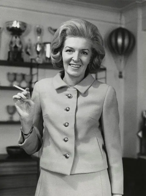 National Portrait Gallery / Godfrey Argent A 1960s-style black-and-white image of the woman in the above picture of the plane. She is wearing a button-up jacket and skirt combination. She is smiling at the camera and holding a lit cigarette in her right hand.