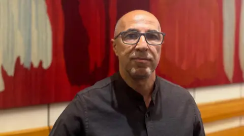A bald, middle aged man wearing a black shirt and glasses. There is a large colourful art work on the wall behind him. 