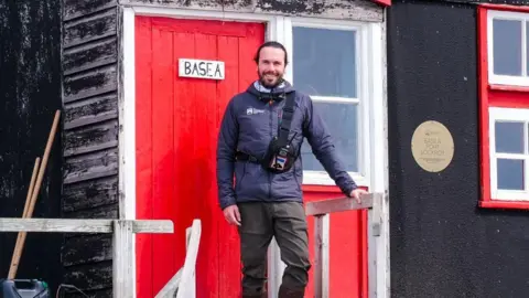 Tiago Figueiredo/UKAHT Pete Watson stands outside a wooden cabin with a red door. He is smiling, wearing a coat, trousers and scarf.