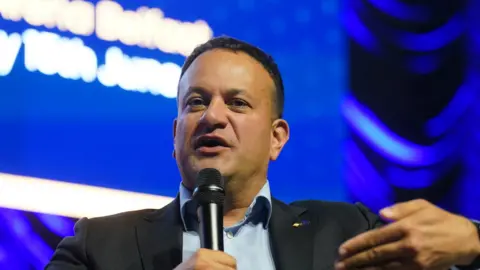 Leo Varadkar is speaking into a microphone that he is holding in his hand. He is wearing a light blue shirt and black blazer. Behind him is a large blue background with bright, white writing on it. 