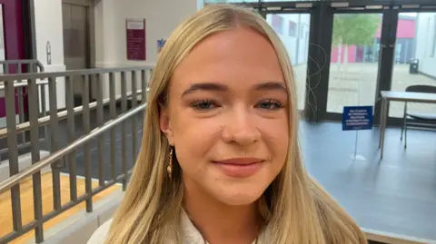 A girl, Rebecca McVeigh. She has long blonde hair and is wearing gold earrings. She is pictured in her school. 