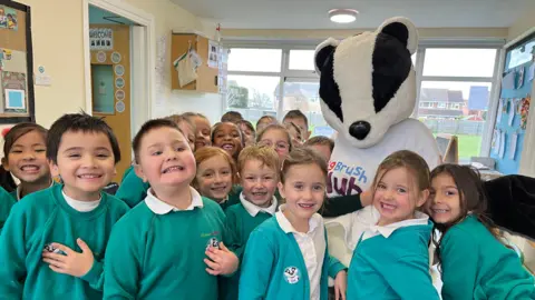 Children in a classroom showing their teeth to the camera, wearing teal-coloured sweatshirts. An adult in a badger costume is also looking at the camera with the word brush on their shirt