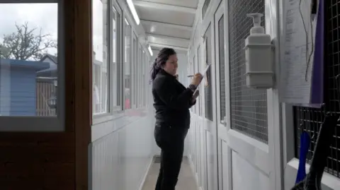 Ginny Parratt A female volunteer holding a clipboard and a pen standing in a corridor with several doors leading to individual rooms housing the animals. There is a bottle of hand sanitiser on the wall in the foreground and outside the window is a blue shed and wooden fence