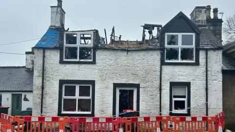 BBC A house in New Abbey which has been hit by fire. The roof has caved in and there are red and white barriers, and police tape, surrounding the building. 