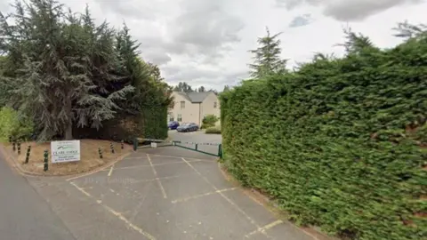 Google A section of a cream coloured building with a grey roof can be seen in the gap between two connifer bushes. There is a concrete driveway leading up to the house with yellow hatching in front of a small metal gate. 