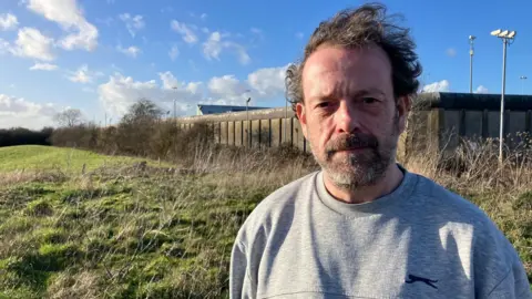 Spike Hughes stands in sunshine in a field next to HMP Long Lartin. The outer prison wall is visible in the background as well as floodlights. He is wearing a grey jumper.