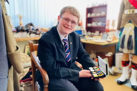 Boy, 16, smiling, wearing glasses, black jacket, white shirt, red white and blue striped tie, holding medals, with antiques in the background.