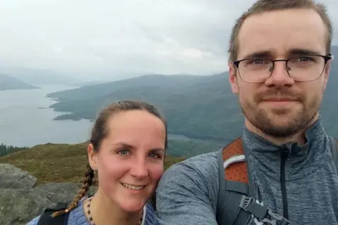 Hunter family A woman and man pictured on top of a hill, taking a selfie. The woman has long brown braided hair, the man has short fair hair, a beard and glasses
