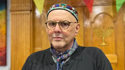 Rabbi René Pfertzel is stood in front of a wooden case and is wearing a neck tie and a yarmulkes as he stands in the synagogue.