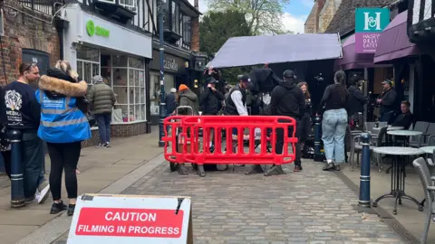 Holly Nichols/BBC A cobbled street in Hitchin where a film crew are filming a drama. On either side are shops, on the right there are tables and chairs on the pavement. In the middle of the street is a red barrier with the film crew. A mix of men and women are milling around, fiddling with cameras. On the left foreground is a sign resting on the ground, saying CAUTION FILMING IN PROGRESS.