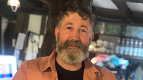 A middle-aged man with curly black hair and a grey beard. He is looking into the camera and is wearing a black t-shirt with orange shirt over the top. He is stood in a room with bottles and cups on shelving behind him, with a low beamed ceiling.