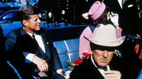 Kennedy seen in the backseat of an open convertible seated next to his wife who is wearing a pink outfit. A man in front of him is wearing a white cowboy hat