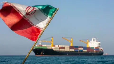 An tattered Iranian flag billowing against a blue sky with the Strait of Hormuz and a large trading ship in the background in soft focus