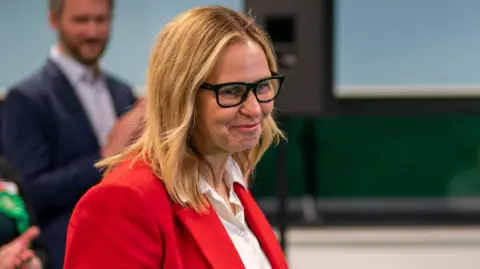 Helen Godwin is walking across the frame in a red suit jacket. She is smiling and looks happy. People in the background are clapping.