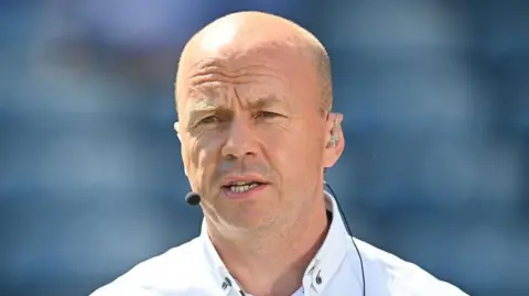 Canavan is bald and is wearing a white shirt and has a mic on.