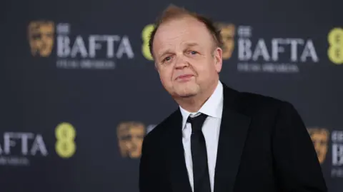 Reuters Toby Jones wearing a white shirt, black tie and black suit jacket, standing on the red carpet at the Baftas. A black board with Bafta branding on it can be seen in the background.