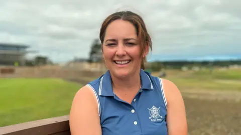Portrait of a person seated on a wooden bench at a golf course, wearing a blue sleeveless polo shirt. Green grass, pathways, and a cloudy sky form a blurred background behind the subject.