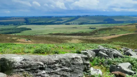 Duncan Hutt Large stones in the foreground but beyond that is a view of fields, hedgerows and hills with just one or two farms visible